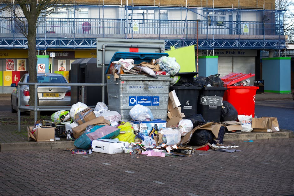 A large collection of overflowing rubbish bags and assorted waste items, including black plastic bags, cardboard boxes, and paper waste, are piled up on a paved sidewalk in front of a commercial area. The waste is situated near a metal railing, with some packaging and loose debris scattered across the ground, indicating a site of irregular disposal. Behind the waste pile, there are parked cars, including a silver vehicle, and a building with signage and storefronts, suggesting an urban retail or shop area. The waste appears to consist of mixed household or commercial rubbish typical of a private or independent rubbish collection service, such as Rubbish Removal Wembley. The scene is set in daylight, with the background featuring some scaffolding or construction elements on the upper part of the building, adding context to a busy, possibly congested urban environment. The overall composition reflects a typical example of unmanaged rubbish accumulation that suitable waste removal services could address in a timely manner, supporting the importance of effective rubbish clearance for maintaining cleanliness in commercial locations.