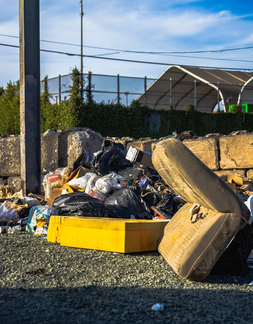 A roadside collection of refuse consisting of black and transparent plastic rubbish bags, discarded cardboard boxes, and a large rubber tyre leaning against a stone wall. The debris is spread across a gravel surface, with some of the packaging torn open and contents spilling partially onto the ground. In the background, there is a stone wall and a metal fence, with trees and shrubbery visible behind it. Above, the sky is bright with some wispy clouds, and overhead power lines stretch across the scene. To the right, part of a green tent or shelter structure can be seen. The scene appears to be outdoors on a paved or gravel area adjacent to a public or private property, illustrating an example of waste accumulation that might require professional rubbish removal services such as those provided by Rubbish Removal Wembley, especially for alternative waste handling or on-site clearance options.