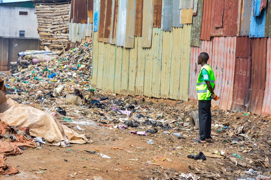 A man wearing a high-visibility yellow vest stands with his hands behind his back on a dirt ground covered with scattered waste materials, including plastic bags, bottles, and other refuse. He is positioned to the right of the image, facing a tall, makeshift wooden wall constructed from unevenly painted and weathered wooden planks in shades of brown, green, blue, and reddish tones. To the left and in the background, there are piles of additional debris, possibly discarded packaging and organic matter, with a rough, unpaved surface showing patches of exposed soil and loose trash. The environment appears to be an informal rubbish dumping area, with no visible waste containers or recycling bins, indicating a site likely managed through private waste collection services, aligning with independent rubbish removal practices often necessary in urban or peri-urban settings. The scene is lit by natural daylight, emphasizing the textures of the clutter, the rugged surroundings, and the weathered surface of the wooden wall, illustrating a typical context for private waste clearance or rubbish removal operations.