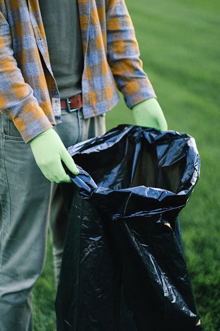 A roadside collection of refuse consisting of black and transparent plastic rubbish bags, discarded cardboard boxes, and a large rubber tyre leaning against a stone wall. The debris is spread across a gravel surface, with some of the packaging torn open and contents spilling partially onto the ground. In the background, there is a stone wall and a metal fence, with trees and shrubbery visible behind it. Above, the sky is bright with some wispy clouds, and overhead power lines stretch across the scene. To the right, part of a green tent or shelter structure can be seen. The scene appears to be outdoors on a paved or gravel area adjacent to a public or private property, illustrating an example of waste accumulation that might require professional rubbish removal services such as those provided by Rubbish Removal Wembley, especially for alternative waste handling or on-site clearance options.