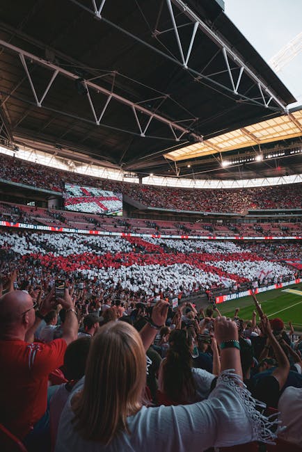 A large crowd of spectators fills the stands inside a modern stadium, with some individuals visible in the foreground using mobile phones to capture moments. The seating area is decorated with a display of thousands of white and red panels, forming a pattern or image, likely supporting a team or event. The stadium features a high, open roof structure with steel trusses and beams overhead, allowing natural light to illuminate the interior. The field area is partially visible near the bottom right, showing a green playing surface. The atmosphere appears lively and energetic, typical of a major sporting event. The scene reflects a setting where spectators are engaged, and the environment highlights the scale and design of the venue, which could relate to alternative ways of hosting large public gatherings or events outside traditional venues, similar to private or independent arrangements in large-scale entertainment or sports venues. Rubbish Removal Wembley might handle waste from such large-event settings, ensuring proper clearance of crowd-generated rubbish in accordance with waste management standards.
