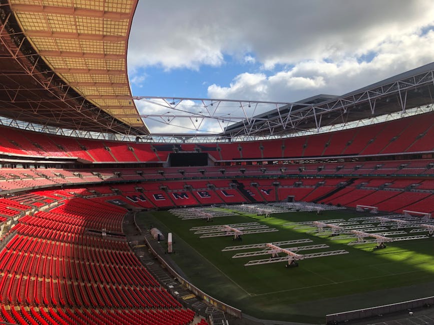 A large sports stadium with a partially covered roof structure overhead. The seating area is primarily red, arranged in circular tiers around the central playing field. The word 'WEMBLEY' is spelled out in large black letters across the middle section of the seats. The grass pitch is green and neatly maintained, with multiple white metal scaffolding structures and equipment positioned on it, likely for event setup or maintenance. The sky above is partly cloudy, with patches of blue visible through the clouds, providing natural daylight that illuminates the entire scene evenly. Surrounding the field, there are some pathways and barriers, with a slight shadow cast by the roof additions, highlighting the stadium's open-air design. The overall environment appears clean and organized, with clear evidence of ongoing preparations typical for a large-scale sporting or entertainment event. This scene relates to activities involving temporary installation or preparation for upcoming events, aligning with independent or private venue management of event-related waste or equipment handling, as would be managed by services like Rubbish Removal Wembley.